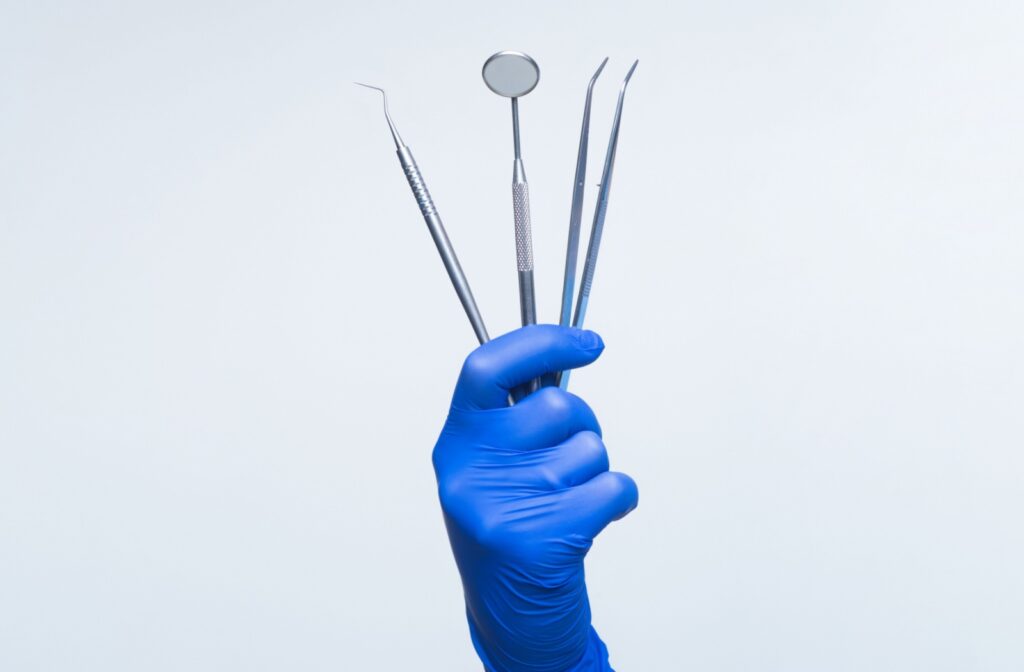 A hand in a blue glove holding up 4 long, thin, metal dental instruments.