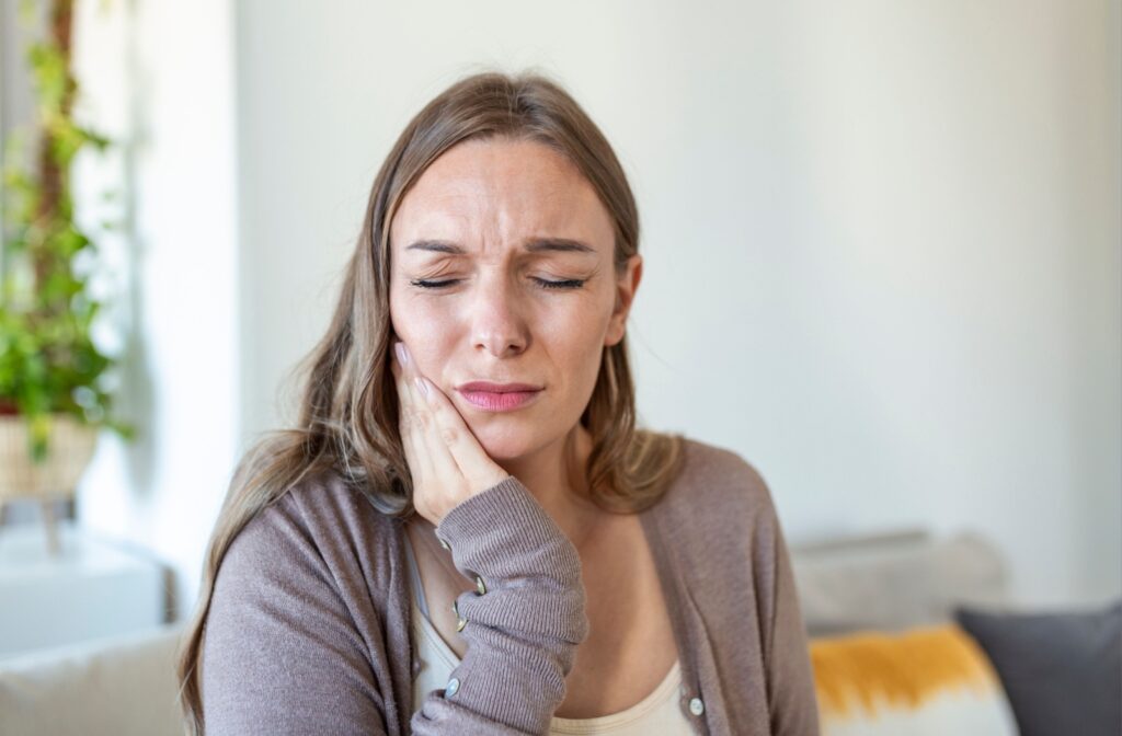 Young woman sitting on a couch holding her cheek in pain, showing discomfort from a toothache.
