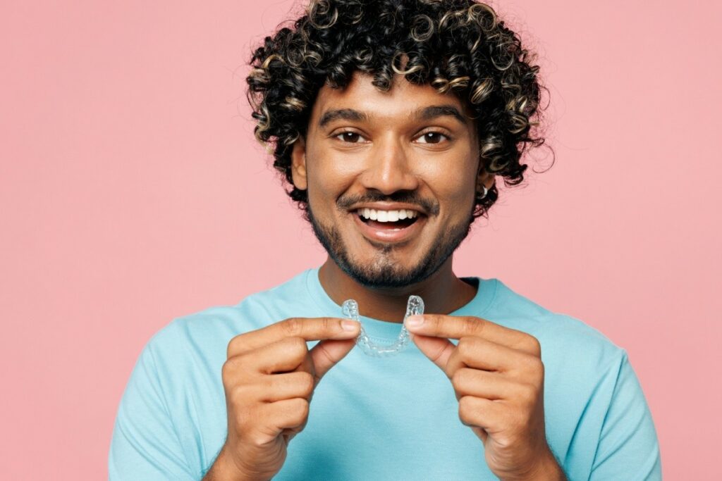 A smiling young man with dark curly hair and a beard holding up a clear dental aligner in front of a solid pink background.