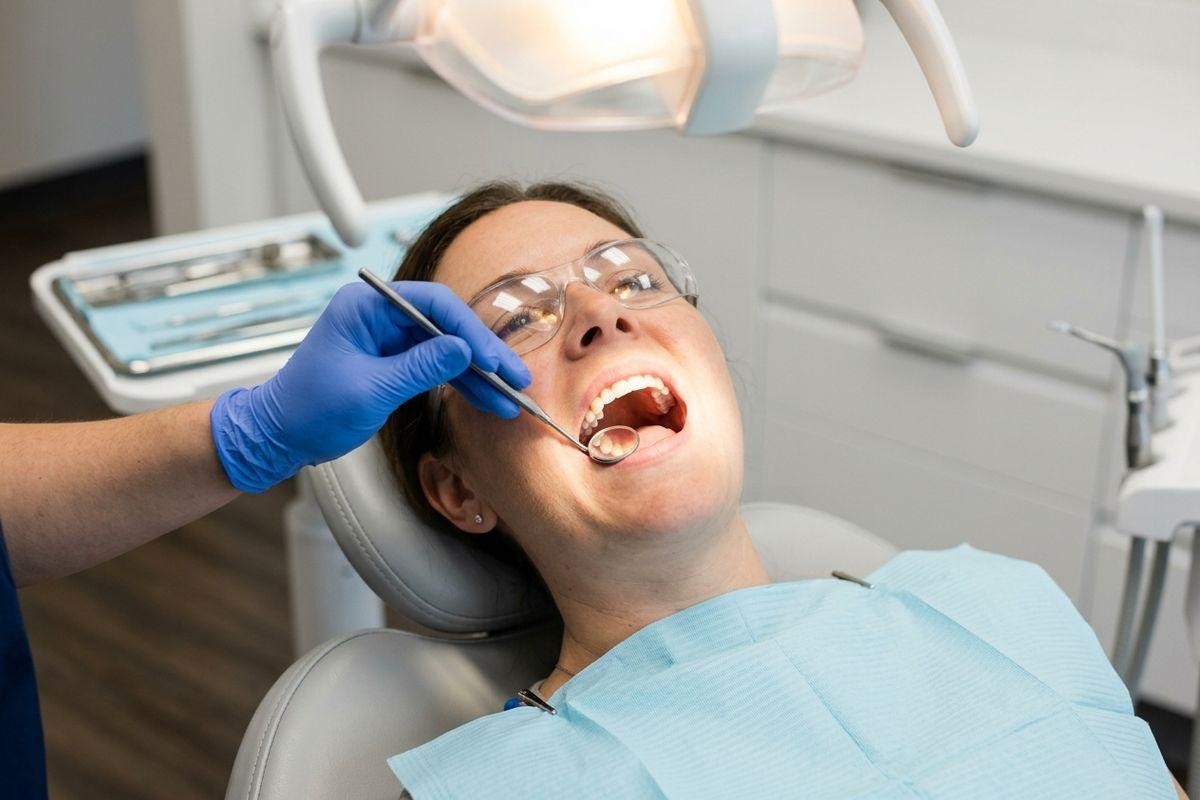 adult-reclined-in-a-dental-chair-with-mouth-open-and-protective-glasses-on-while Adult reclined in a dental chair with mouth open and protective glasses on, while a dentist uses a dental mirror inside their mouth.